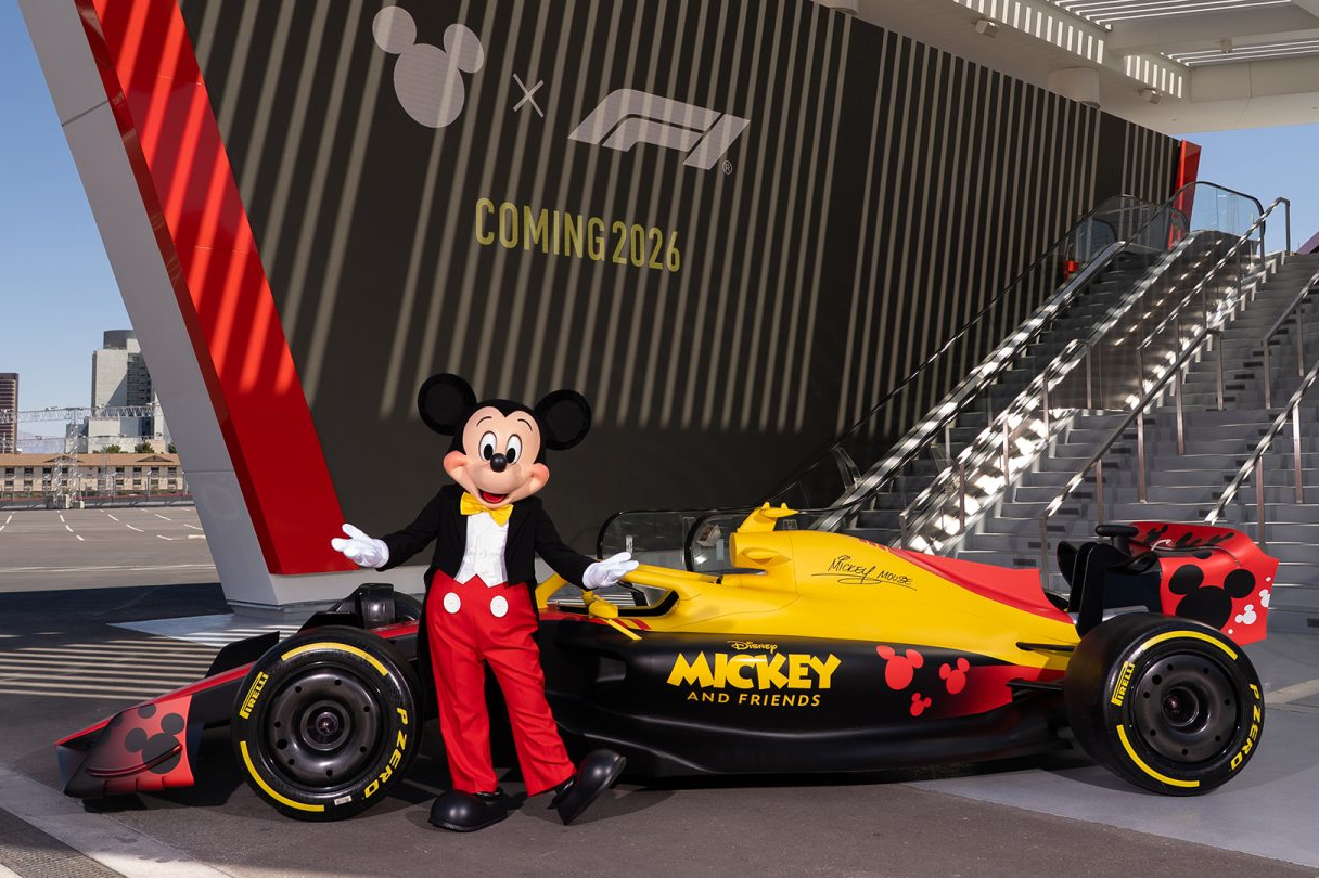 Mickey Mouse stands in front of a Formula One car with a red, black, and yellow Mickey and Friends livery